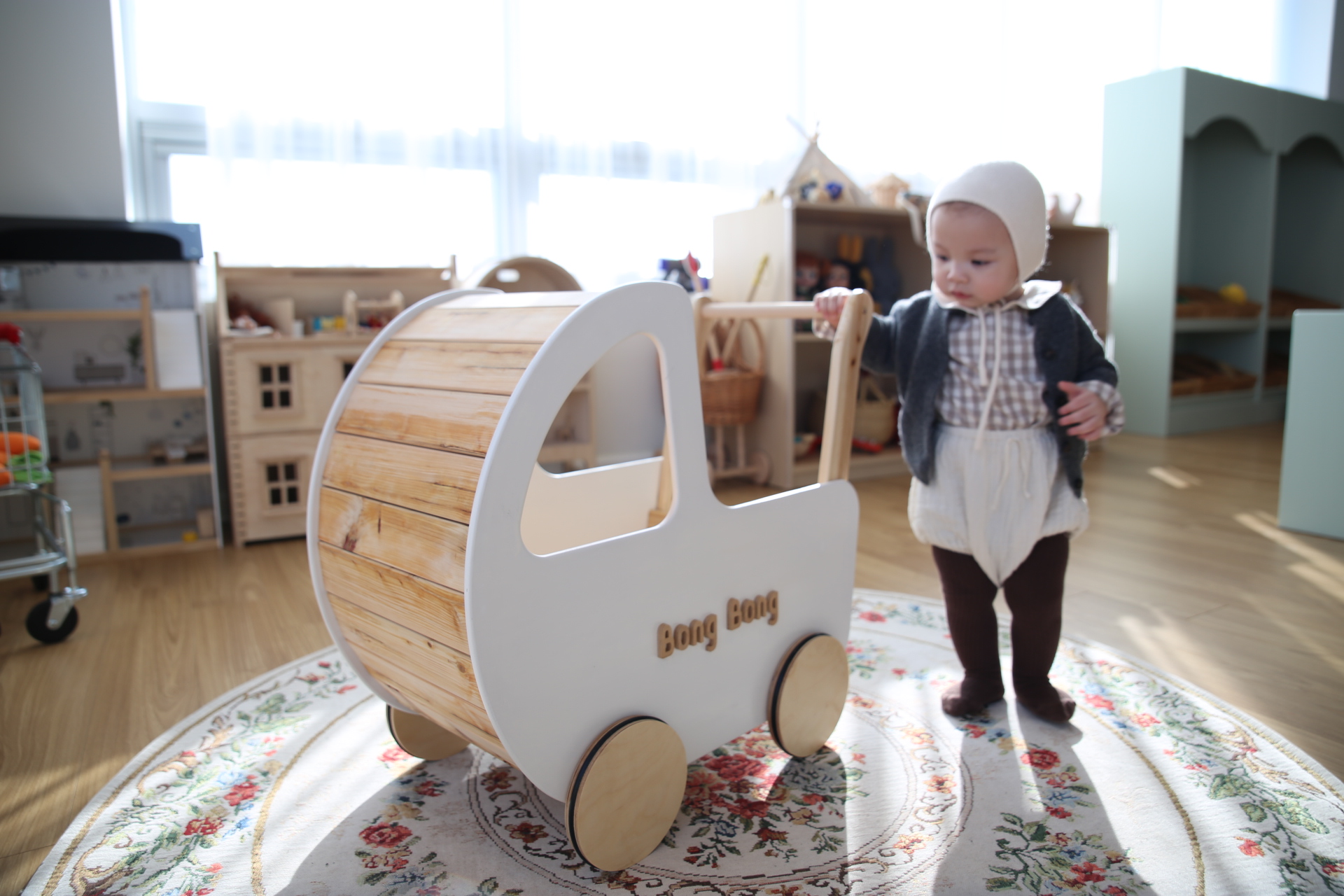 A baby standing and holding the handle of a wooden push walker in a sunlit playroom